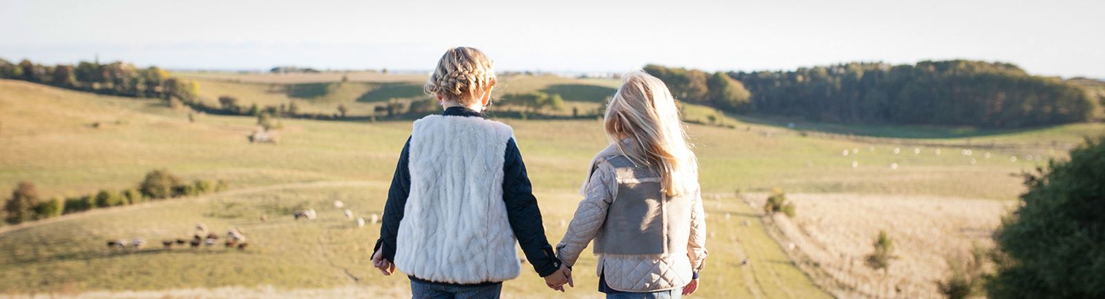 Två flickor står med ryggen emot kameran och håller handen och tittar ut över ett sensommarlandskap