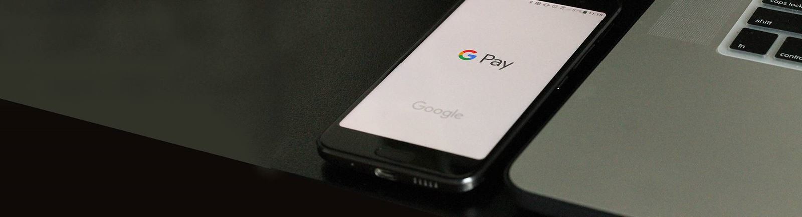 En mobil ligger på bordet sidan om en dator. Mobilen visar Google Pays logga i displayen.