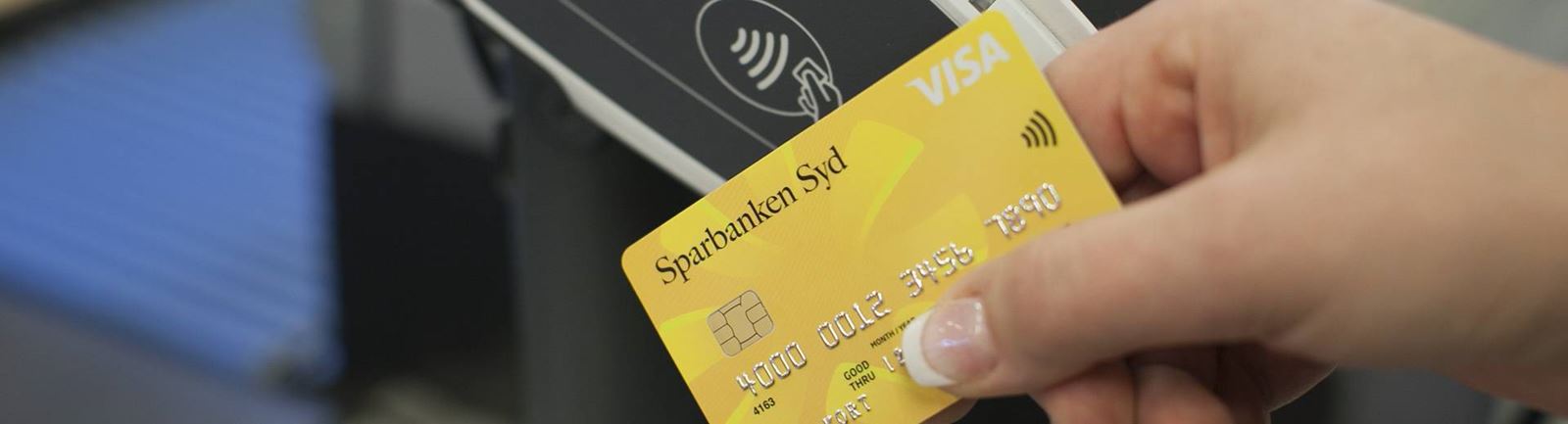 En hand håller ett gult Sparbanken Syd betalkort mot en betalterminal