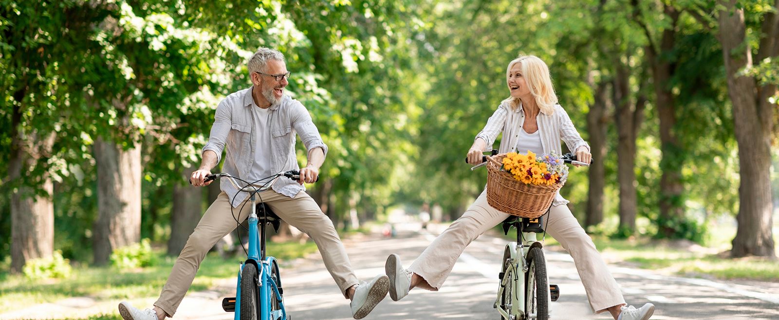 Äldre man och kvinna cyklar med benen ut i luften. Kvinnan har gula blommor i sin cykelkorg
